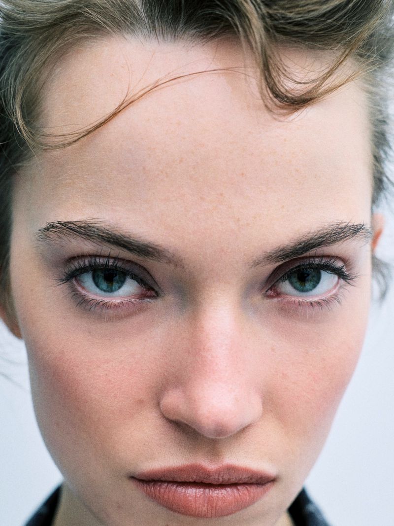 Close-up portrait of a female model with intense gaze and light freckles against a neutral background.