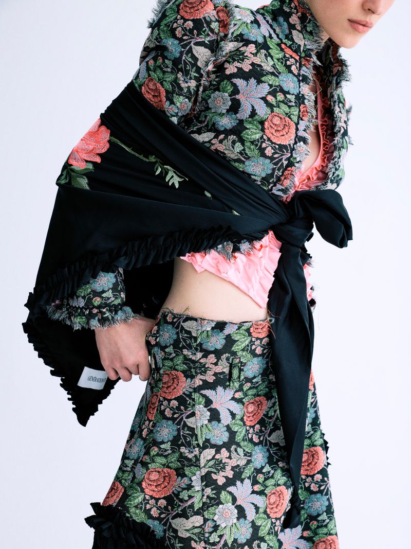 Model wearing a floral print jacket and trousers with a tied black wrap against a white studio background.