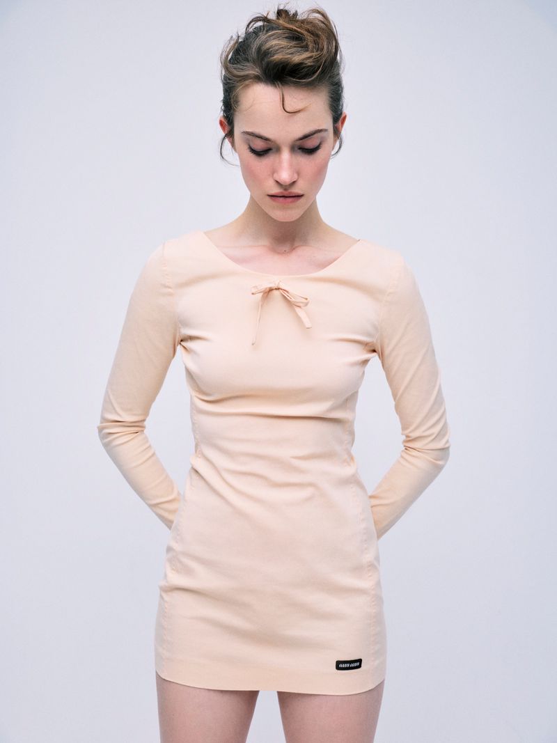 Model wearing a peach long-sleeved mini dress with a small bow detail against a plain light grey background