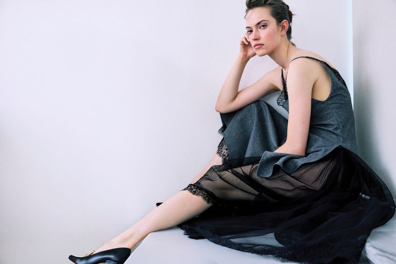 Model seated wearing a grey slip dress with black lace trim and heels against a white seamless background