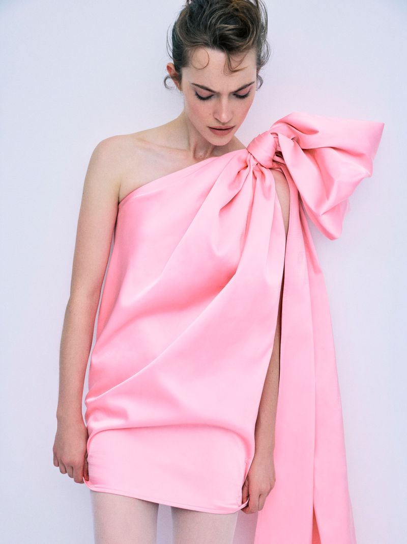 Model Caro Casay in a pink one-shoulder dress with a large bow detail against a neutral studio backdrop