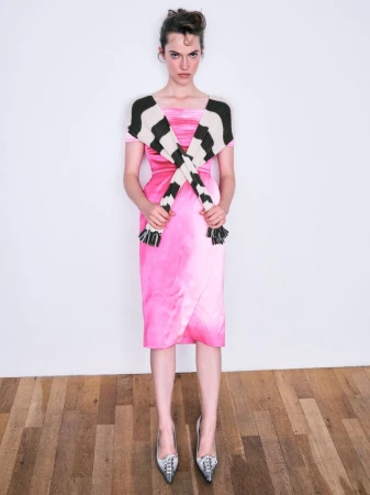 Model wearing a pink dress and patterned scarf standing on wooden flooring against a white background