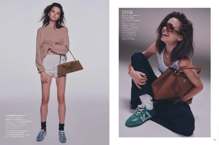 Model wearing a neutral crewneck sweater and white shorts holding a brown leather handbag against a plain grey studio backdrop.