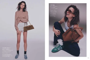 Model wearing a neutral crewneck sweater and white shorts holding a brown leather handbag against a plain grey studio backdrop.