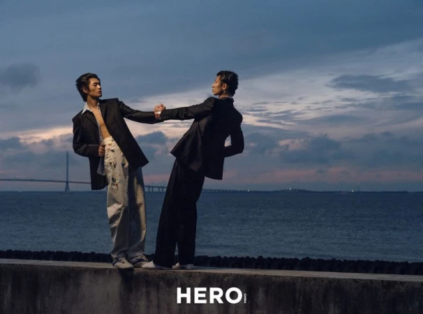 Two male models in dark blazers holding hands on a concrete sea wall under an evening sky, for HERO Magazine.