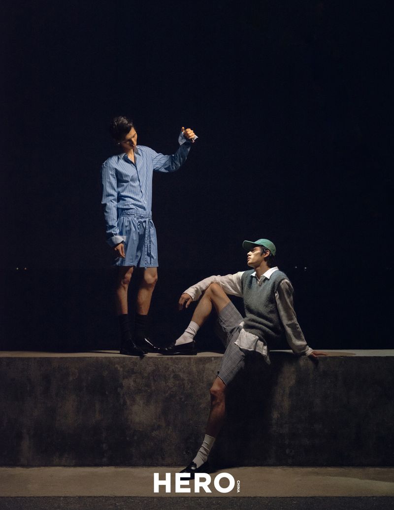 Two male models posing on a low wall at night for HERO Magazine, one standing in blue striped shirt and shorts, one seated in vest and cap