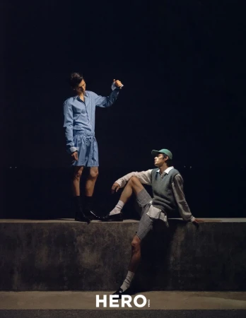 Two male models posing on a low wall at night for HERO Magazine, one standing in blue striped shirt and shorts, one seated in vest and cap