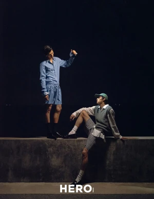 Two male models posing on a low wall at night for HERO Magazine, one standing in blue striped shirt and shorts, one seated in vest and cap
