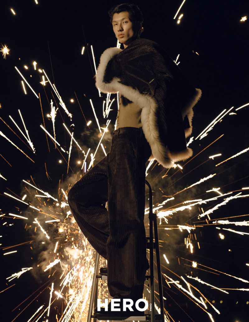 Model standing on a stepladder wearing a dark fur-trimmed coat surrounded by bright flying sparks in the dark.