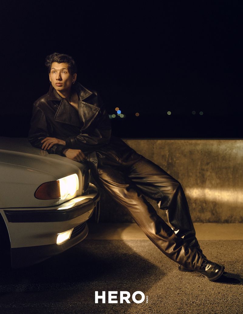 Model wearing a black leather jacket and trousers leaning against a white car at night with distant lights