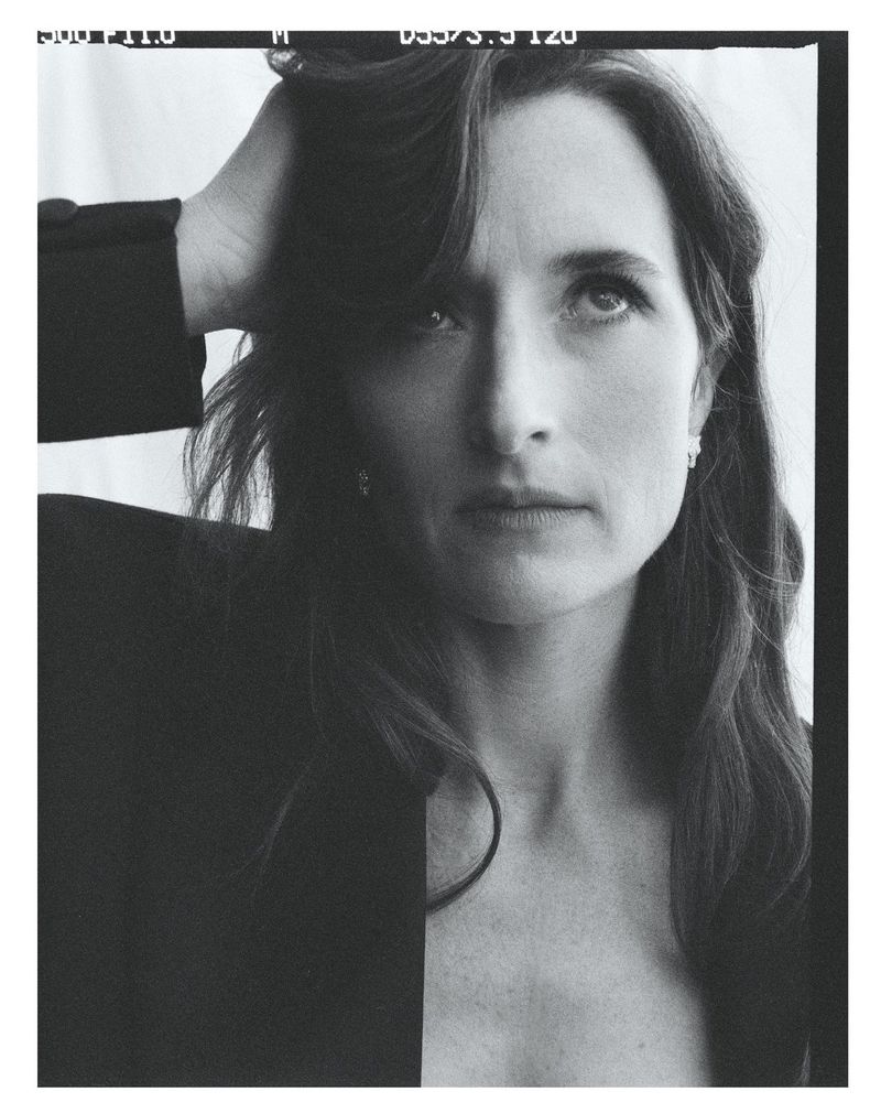 Portrait of Grace Gummer with dark hair running her hand through her hair, wearing a dark blazer against a white backdrop.