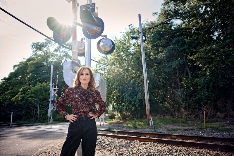 Isabelle Huppert standing by railway tracks wearing a floral print blouse and dark trousers with sunlight shining through signals