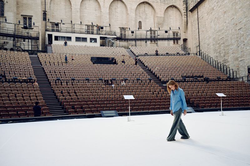 Isabelle Huppert walking across a large empty stage before tiers of empty outdoor theater seating