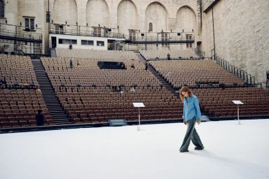 Isabelle Huppert walking across a large empty stage before tiers of empty outdoor theater seating