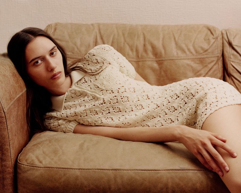 Model lying on a tan leather couch wearing a cream crochet knit polo dress for a Clash Magazine editorial feature.