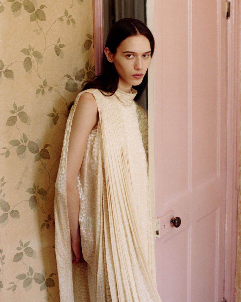 Model in a cream pleated dress standing in a doorway next to vintage floral wallpaper.
