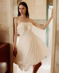 Model wearing a white strapless minidress with oversized bows posing in a doorway for Clash Magazine