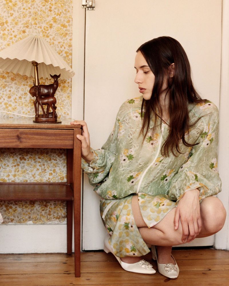 Woman in floral patterned green dress squatting beside a wooden bedside table with a lamp in an interior setting