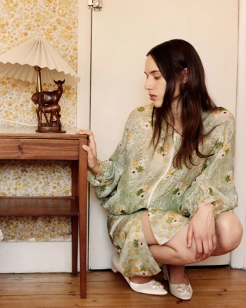 Woman in floral patterned green dress squatting beside a wooden bedside table with a lamp in an interior setting