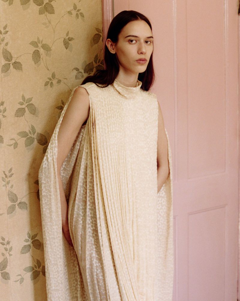 Model in cream sleeveless pleated dress standing against a patterned wallpaper and pink door for Clash Magazine