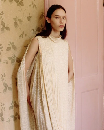 Model in cream sleeveless pleated dress standing against a patterned wallpaper and pink door for Clash Magazine