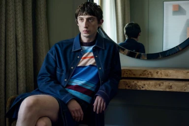 Model wearing a blue denim shirt jacket over a striped sweater in a hotel room with a round mirror on the wall.