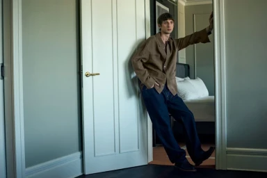 Male model in a brown jacket and blue denim jeans standing in a hotel room doorway, photographed by Roger Rich for Zoo Magazine.