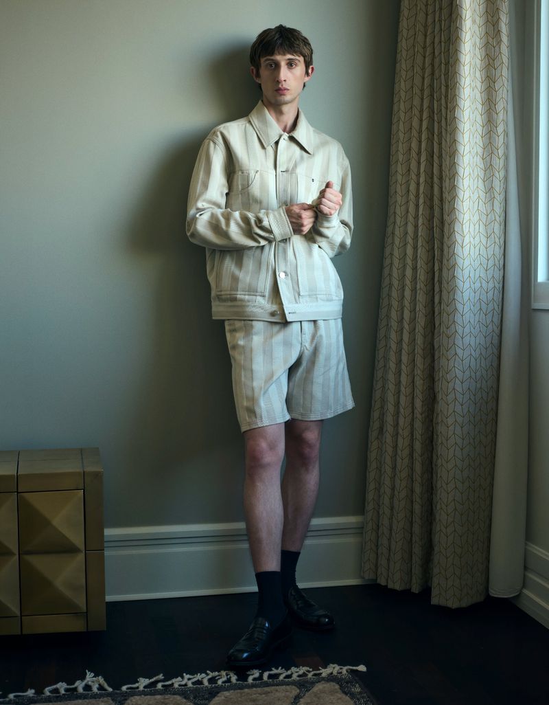 A male model wearing a striped matching jacket and shorts leaning against a plain wall indoors.