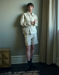 A male model wearing a striped matching jacket and shorts leaning against a plain wall indoors.