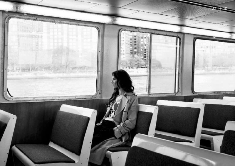 Monochrome shot of woman sitting on a train, gazing out the window, wearing a trench coat, with city buildings visible through the glass.