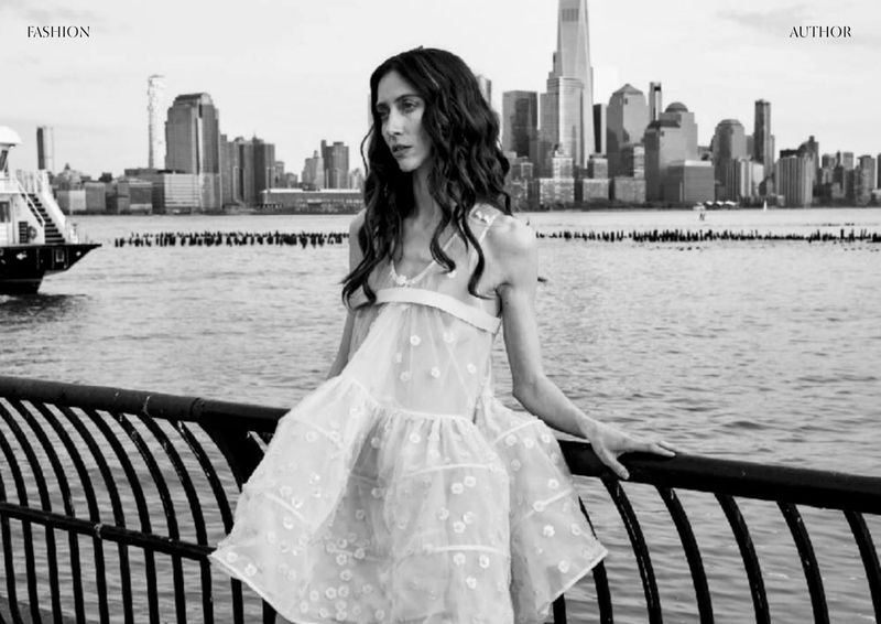A young adult woman poses in a white, tiered dress near a black metal railing, with a water view of New York City skyscrapers behind her.