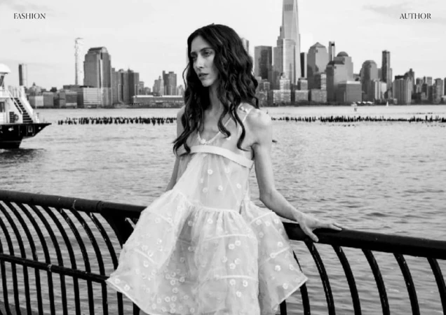 A young adult woman poses in a white, tiered dress near a black metal railing, with a water view of New York City skyscrapers behind her.