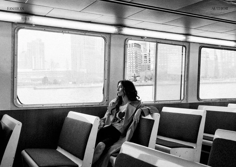 Monochrome image captures a woman in a trench coat sitting on a train, gazing out the window at the city skyline across the water.