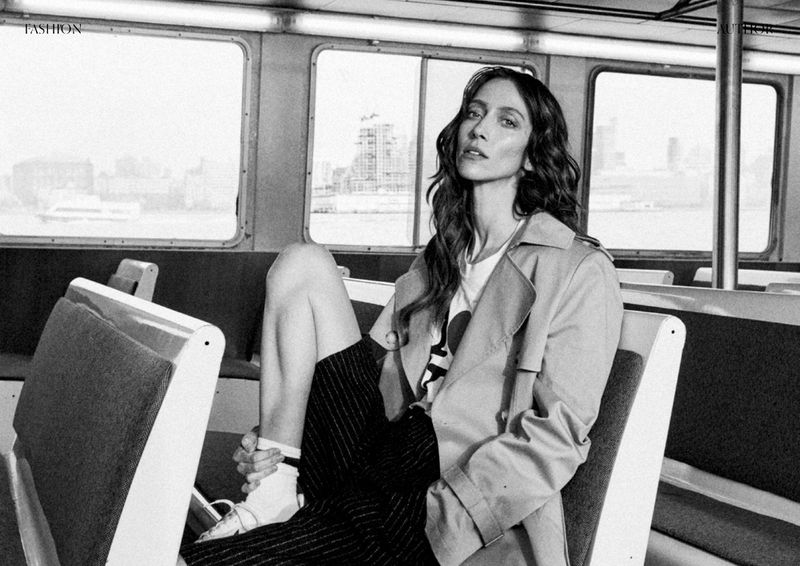 Fashion editorial shot of a woman in a trench coat sitting on a train or ferry, with the city skyline visible through the window.