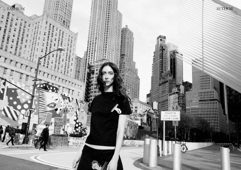 Fashion editorial in black and white featuring a slender woman in an urban environment with tall buildings and unique street art.