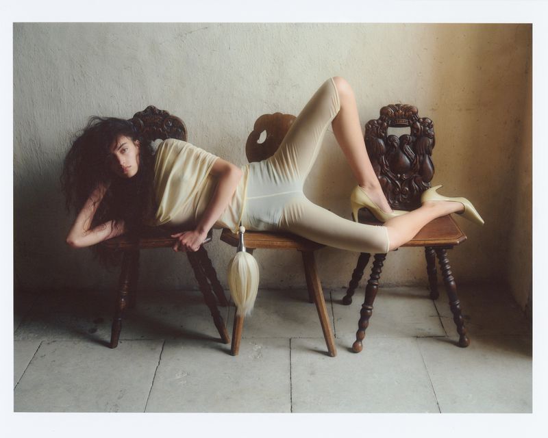 Model lying across three vintage wooden chairs wearing a pale yellow top, leggings, and high heels against a textured stone wall
