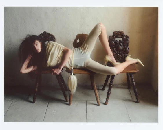 Model lying across three vintage wooden chairs wearing a pale yellow top, leggings, and high heels against a textured stone wall