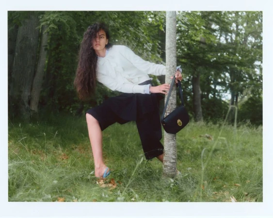 Woman with long dark hair leaning against a tree trunk in a wooded area, wearing a white sweater and dark shorts.