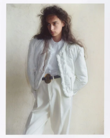 Model with long dark hair wearing a white quilted jacket, white blouse, and white trousers against a neutral wall