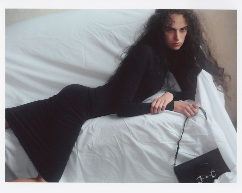 Woman in a black turtleneck dress reclining on a white fabric-covered surface next to a black handbag