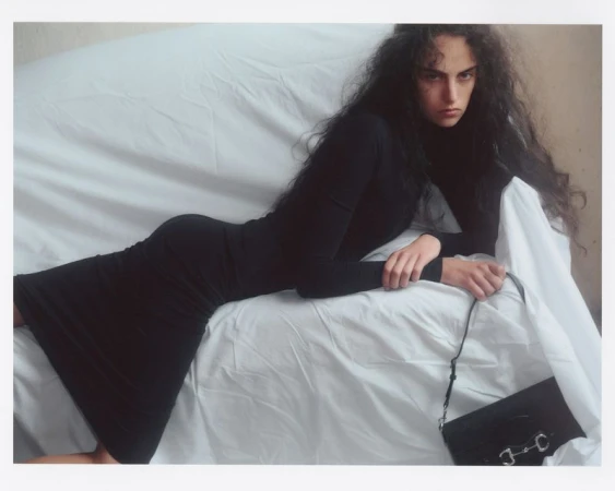Woman in a black turtleneck dress reclining on a white fabric-covered surface next to a black handbag