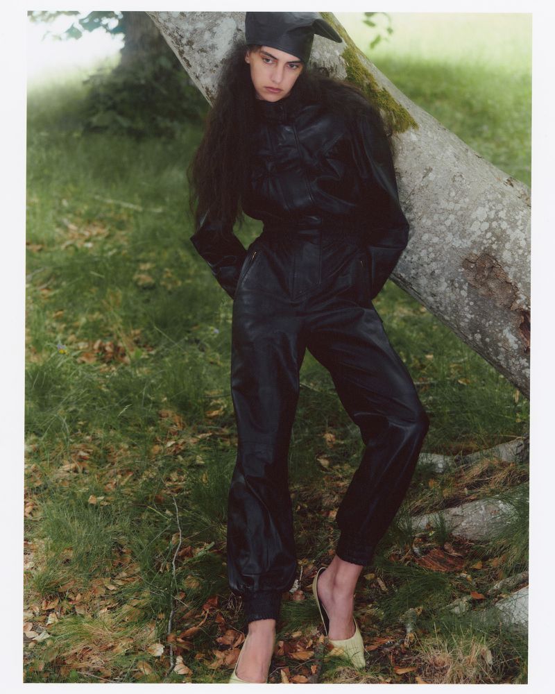 Model in black leather jumpsuit leaning against a large tree trunk outdoors in a grassy woodland setting for Love Want Magazine.