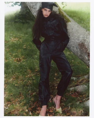 Model in black leather jumpsuit leaning against a large tree trunk outdoors in a grassy woodland setting for Love Want Magazine.