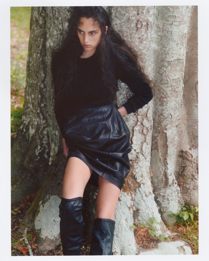 Model with long dark hair leaning against a large tree trunk wearing a black top, leather skirt and boots.