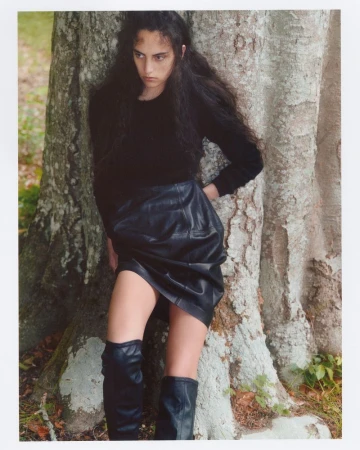 Model with long dark hair leaning against a large tree trunk wearing a black top, leather skirt and boots.