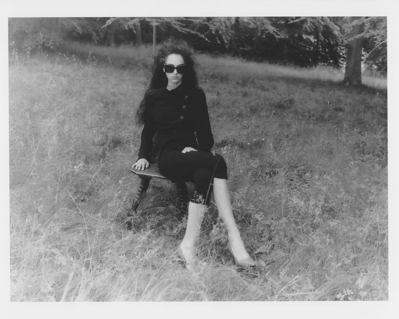 Monochrome fashion portrait of a woman in black clothing and sunglasses seated on a wooden stool outdoors in a grassy field