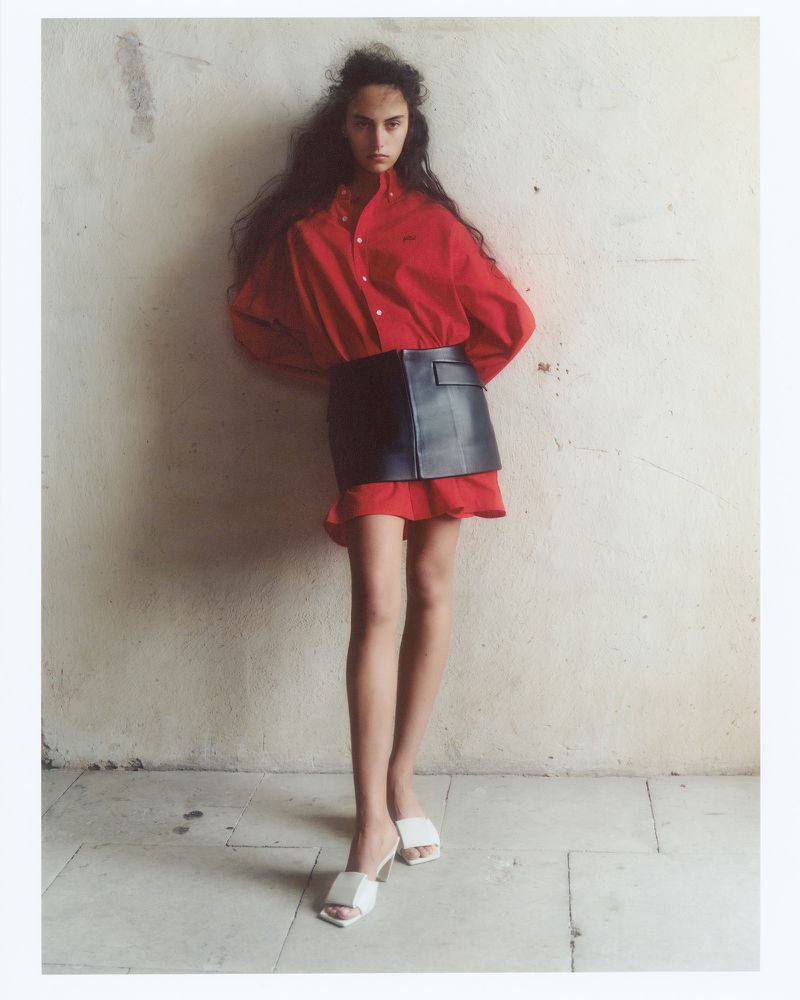 Model wearing a red button-down shirt and black leather skirt standing against a textured off-white wall
