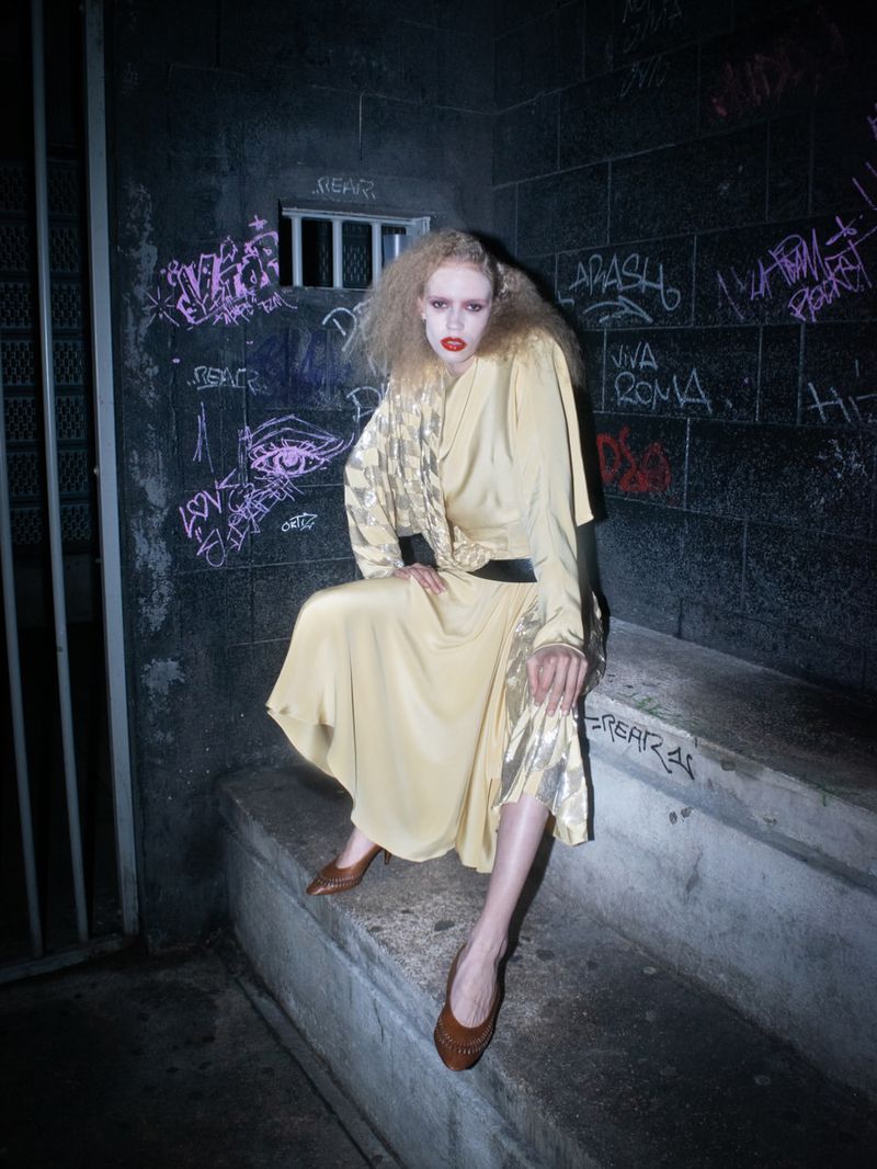 Model with blonde curly hair in yellow dress sitting on concrete steps with graffiti wall background