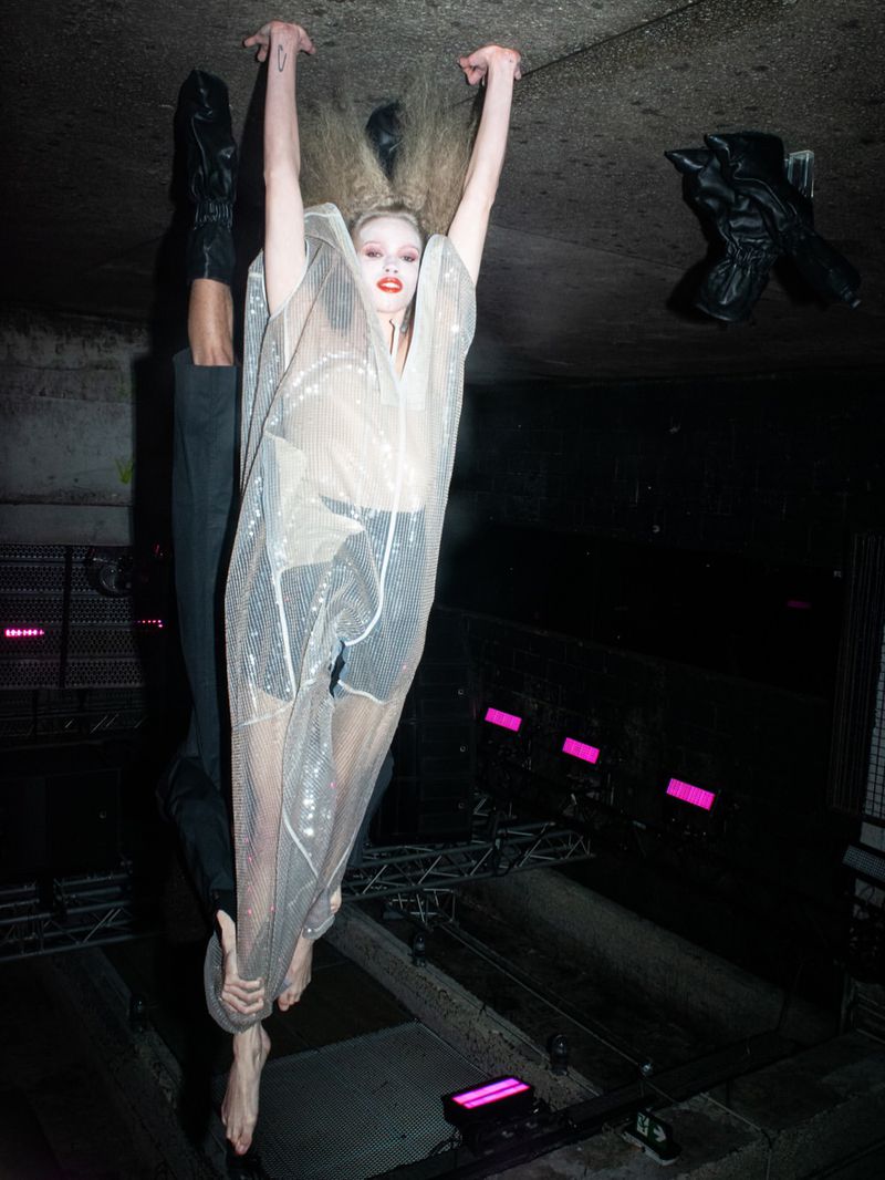 Model hanging upside down from a low ceiling wearing a sheer sequined tunic in a dark subterranean space