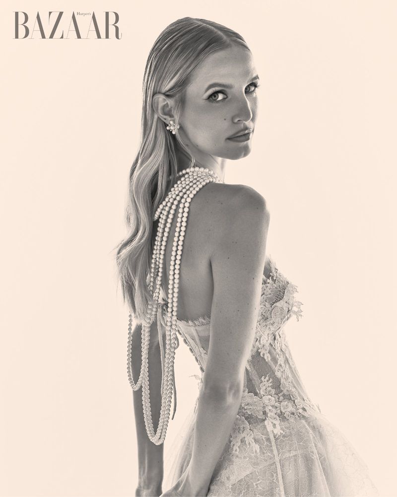 Leonie Hanne in a lace dress with layered pearl necklaces, looking back over her shoulder in a monochrome editorial portrait.